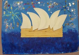 Collage of the Sydney Opera House using white paper plates for the sails and paddle pop sticks for the base. It sits over a dark blue painted ocean. The sky is filled with colourful, sparkly fireworks.