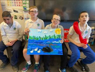 Four boys hold their collage of a feathered black swan, followed by 3 white cygnets, in blue water with green reeds under a blue sky with a yellow sun and white cotton ball clouds.