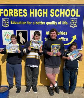 Four students hold their collages, each titled ‘Home’, in front of a school sign that says, ‘Forbes High School, education for a better quality of life.’