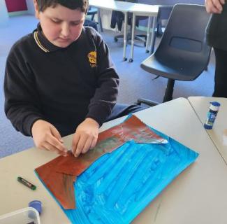 A boy makes a landscape collage. He glues brown paper shapes decorated with green lines on a painted blue canvas.