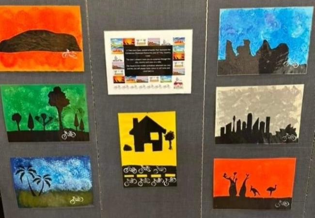 Seven collages depicting various landscapes in silhouette against different coloured backgrounds of red-orange, green, blue, grey and yellow hang on a display board in a square formation as part of the Connections Showcase Submission Exhibition. The house on the yellow background is in the centre.