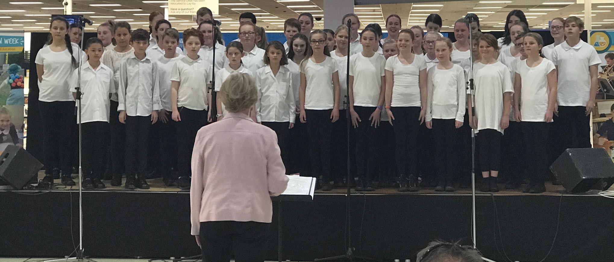 students choir on a portable stage with conductor in front of them.  Students wearing white tshirts and black pants