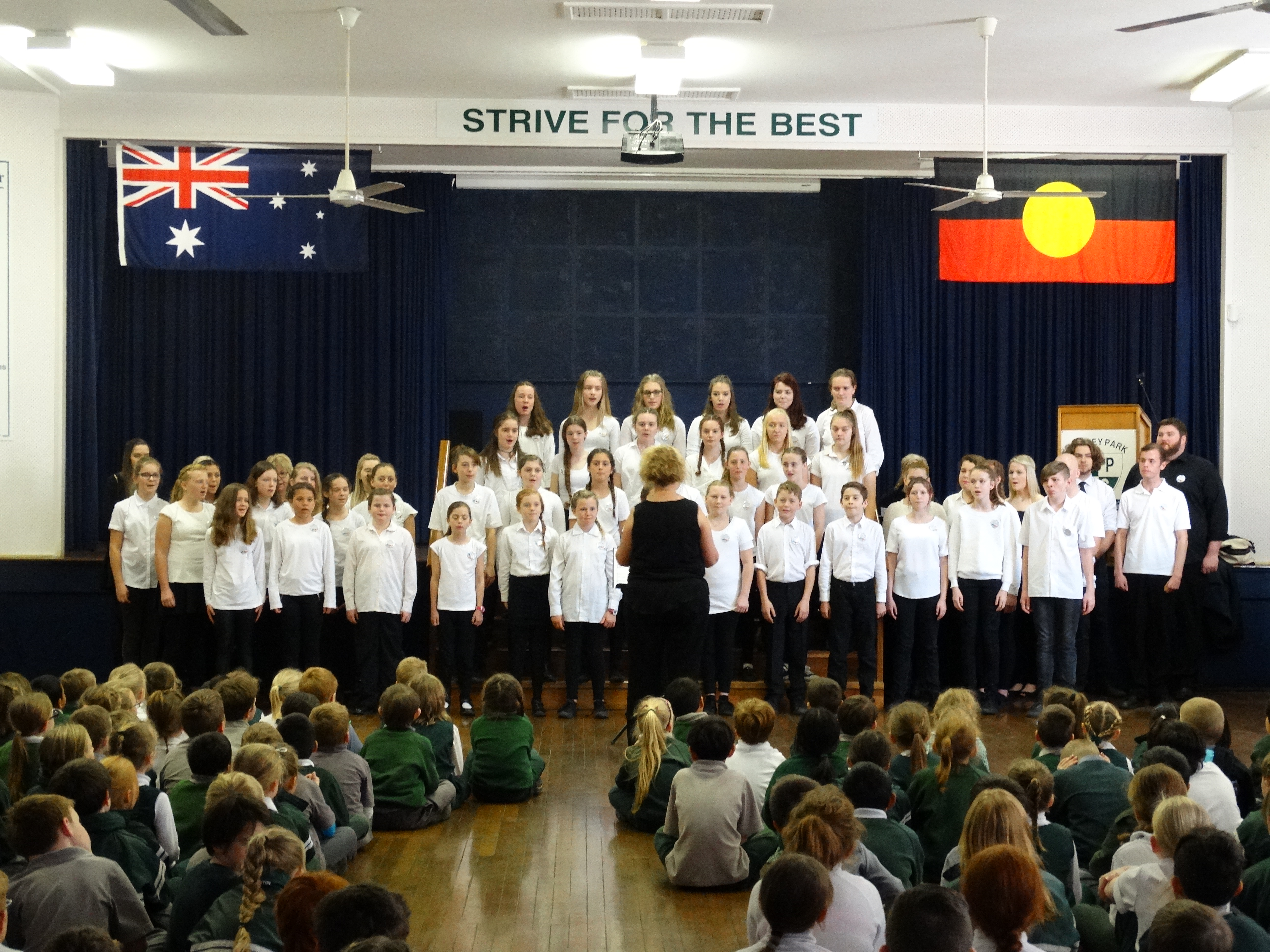 Camp choir performing for a primary school audience wearing white shirts and black pants
