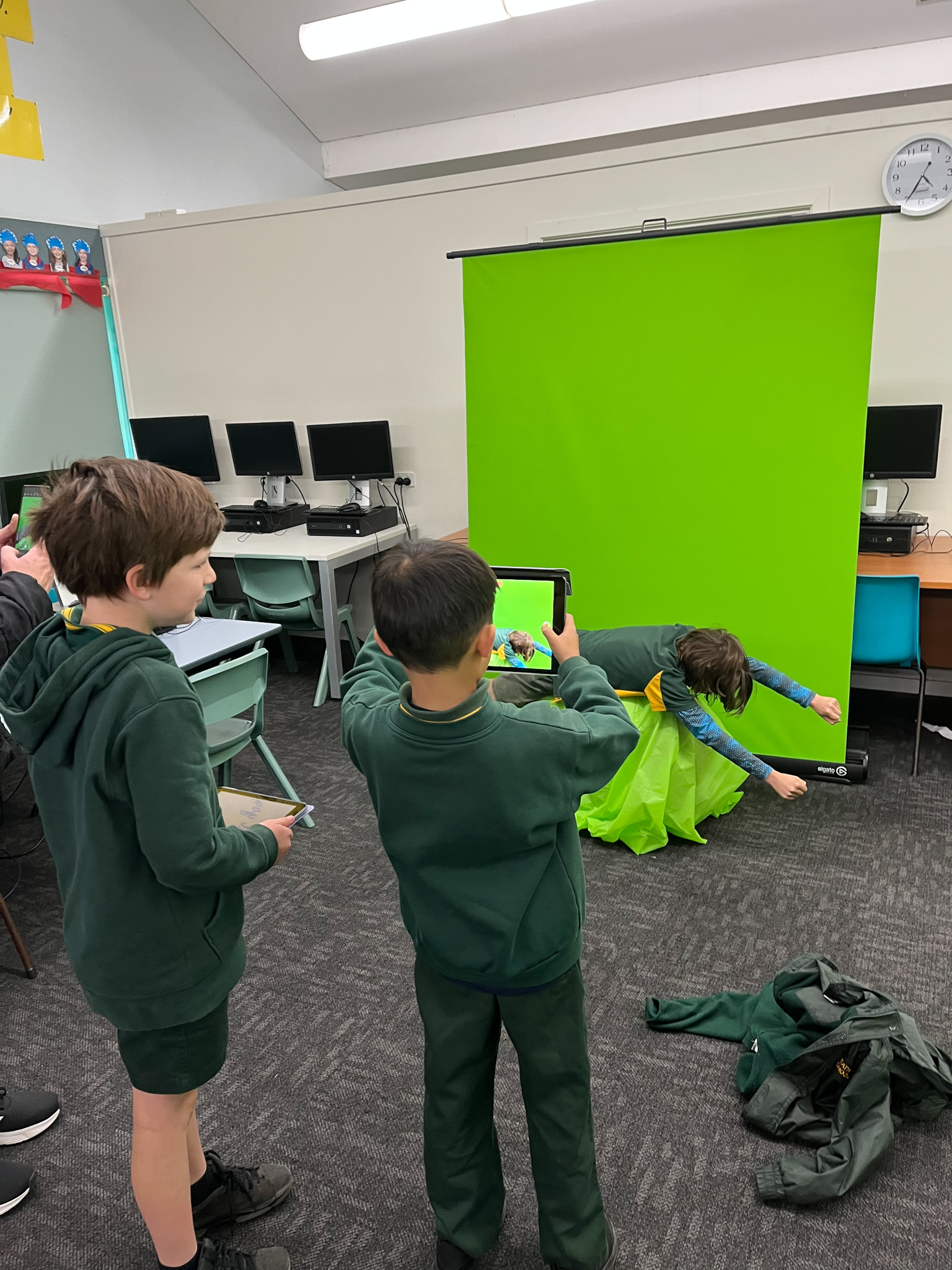 A boy using an iPad to film a scene in front of a green screen. Other students look on.