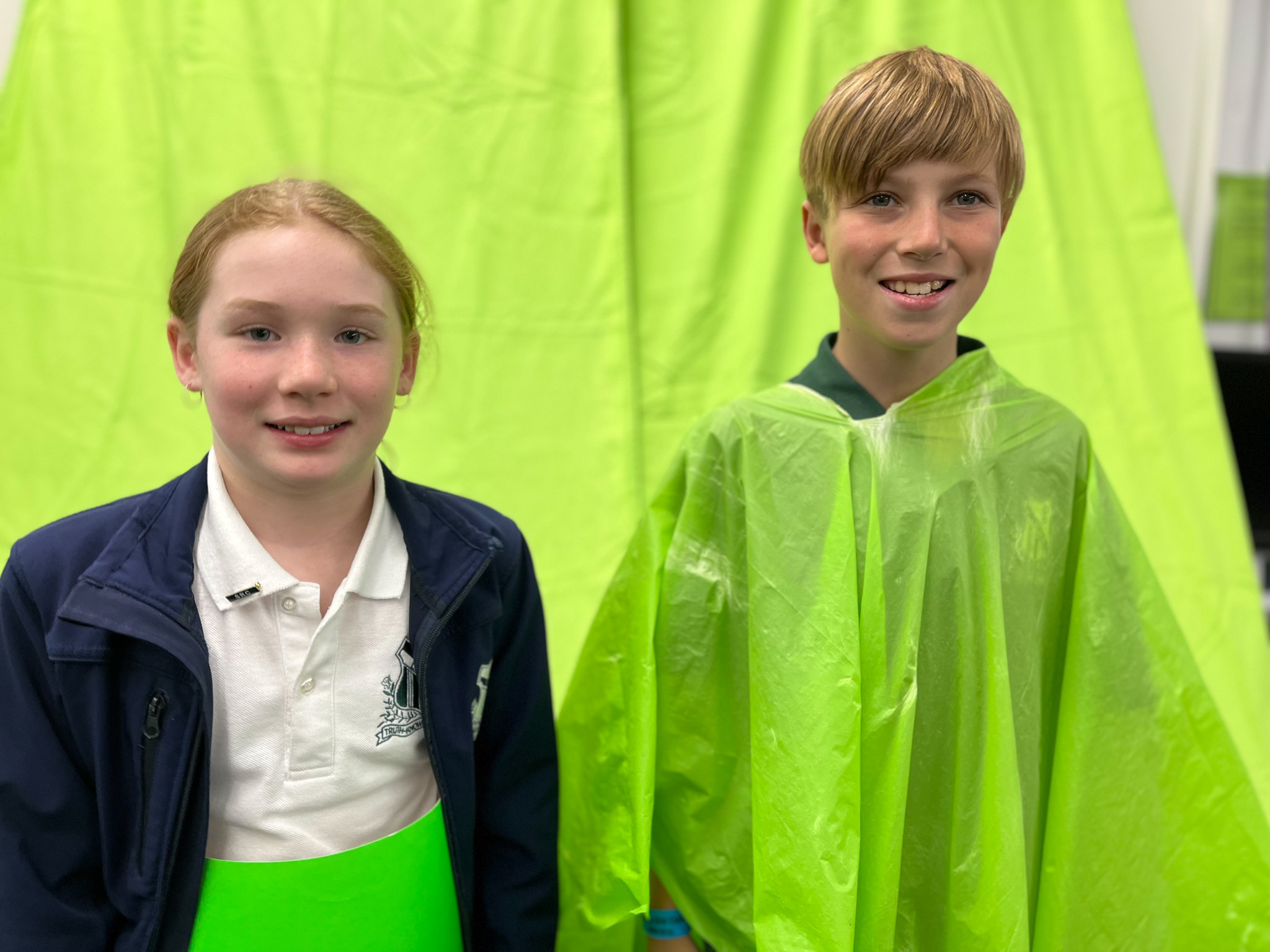 A girl and boy standing in front of a green screen, smiling at the camera. 