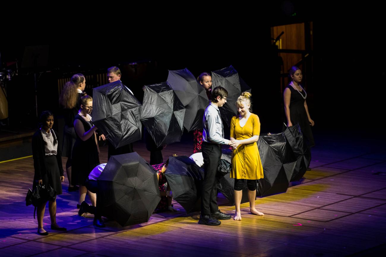 group of students on stage hidden behind black umbrellas with 2 performers in front of them spotlighted