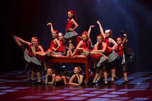 Ensemble of primary dancers on stage in racing style costumes of red, black and white with centre girl standing on a table.