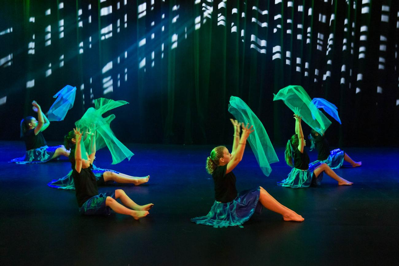 4 students sitting on floor throwing green material into the air on stage