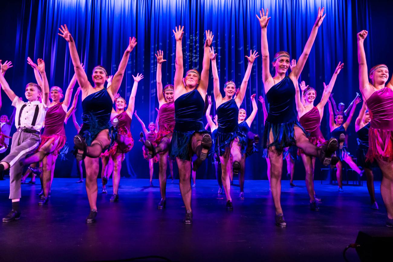 large group of student dancers on stage in flapper style dresses kicking one leg up with arms stretched up