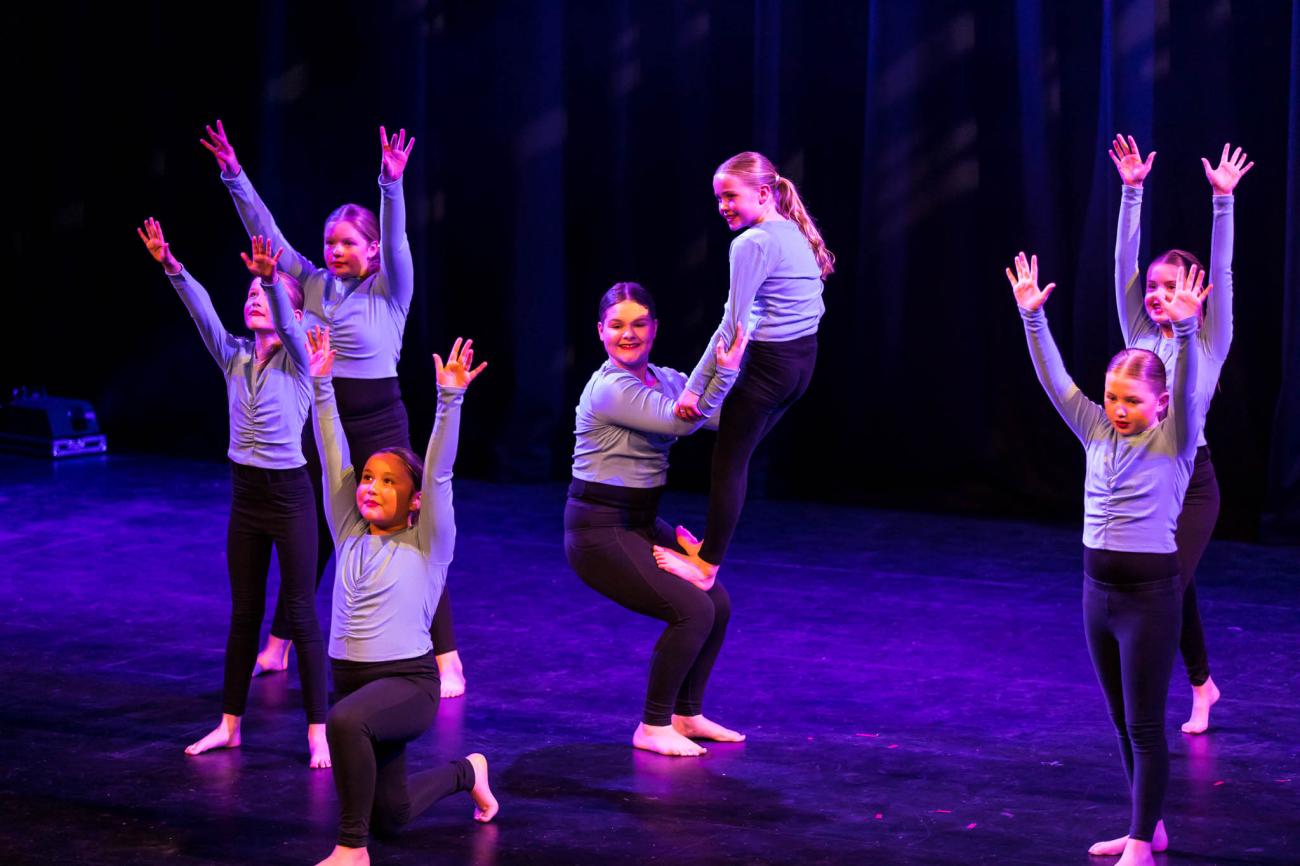 students on stage in purple tops and black pants, 2 students in centre doing a balance, 5 students around them with arms in air