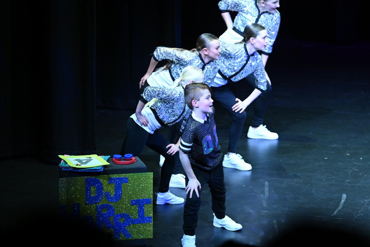 students in a line on stage, boy at front dressed as DJ, students behind in silver
