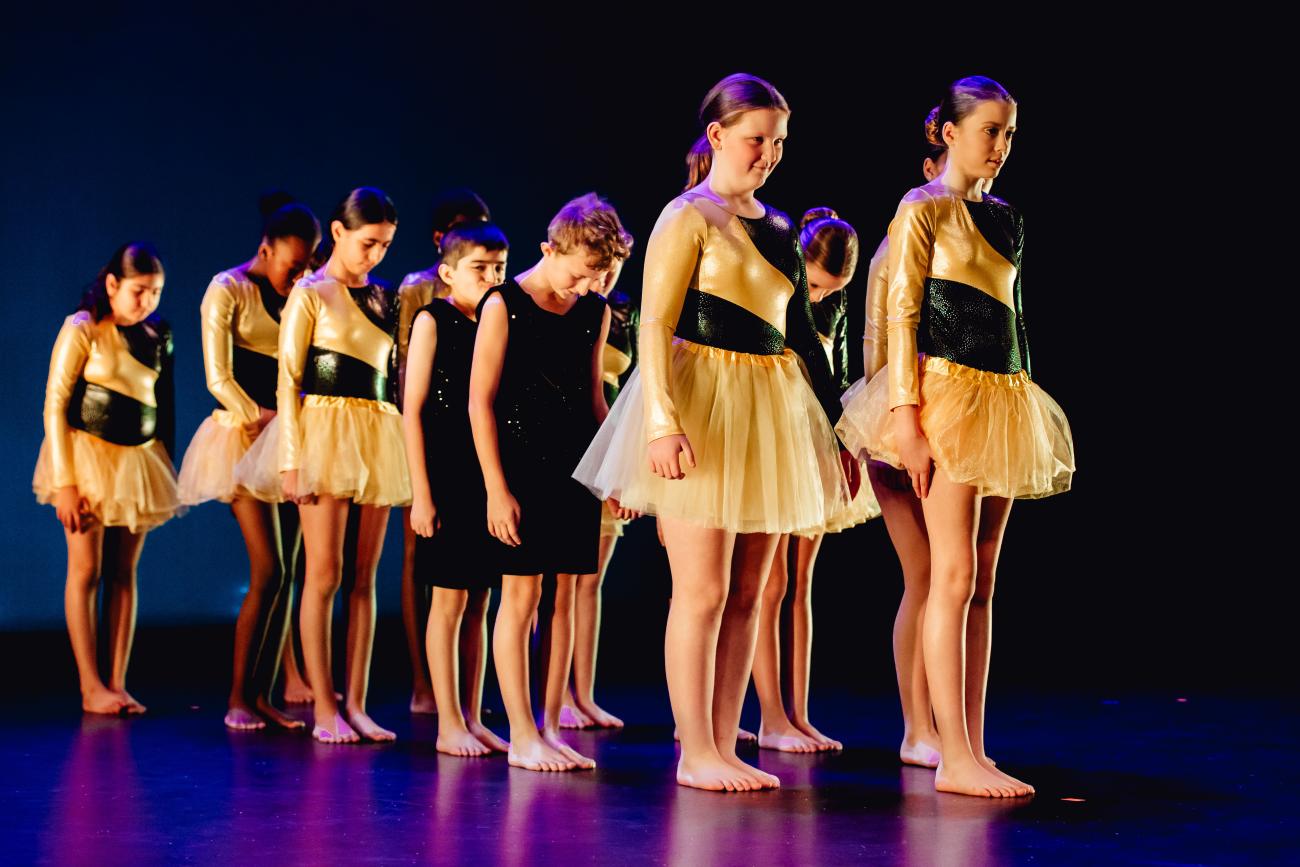 students in 2 lines on stage dressed in yellow and black with arms by side and heads down