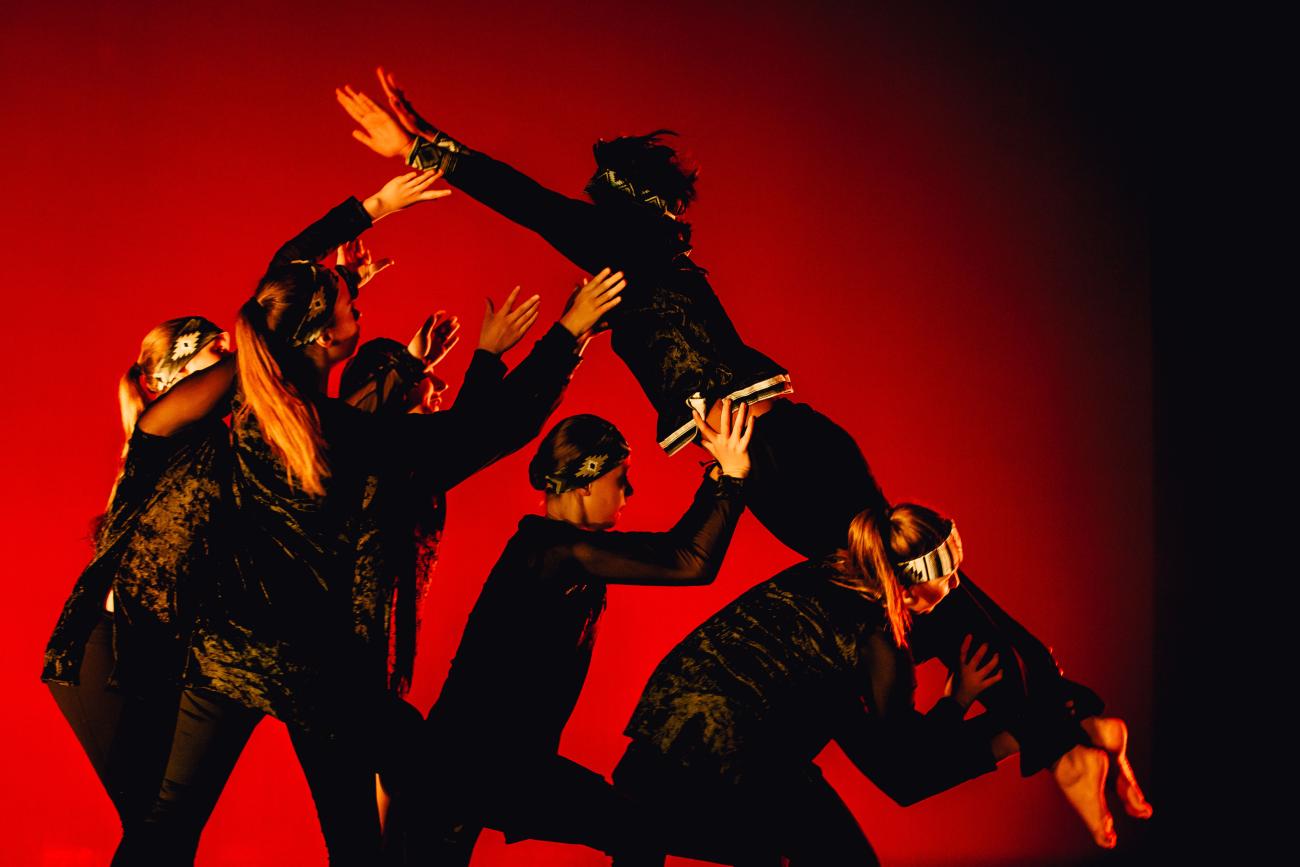 5 students lifting another student into the air dressed in black costumes with red lighting
