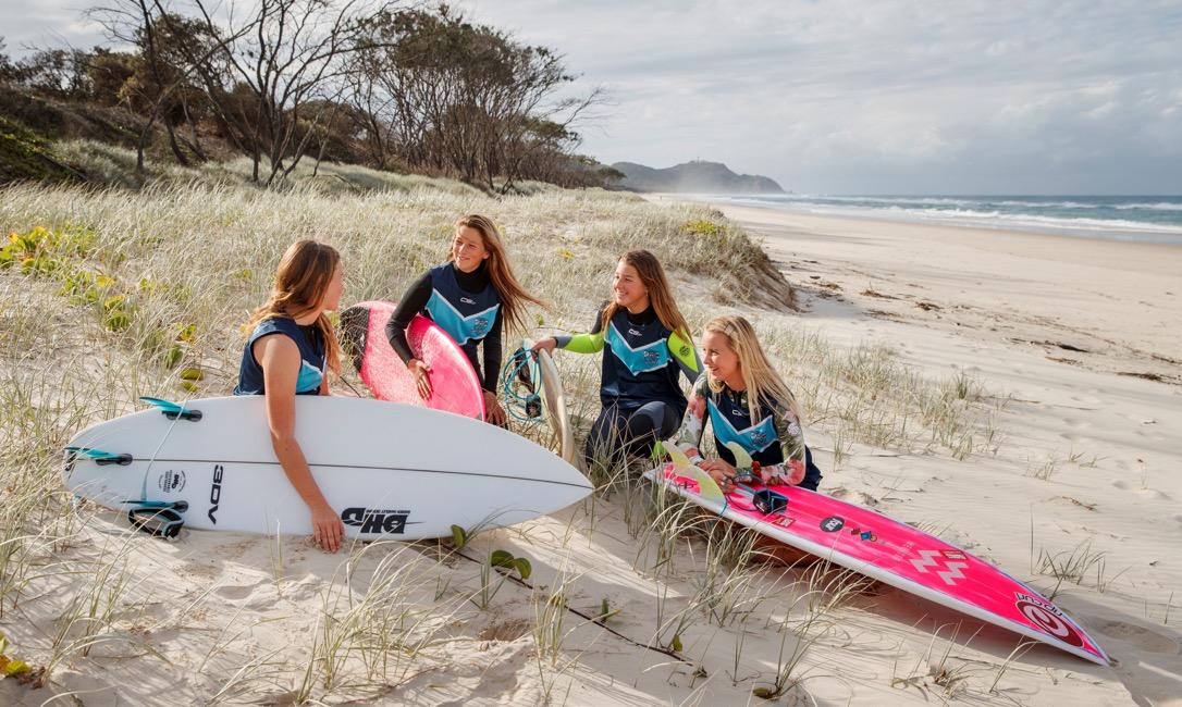 Four students in wet suits holding surf boards at a beach