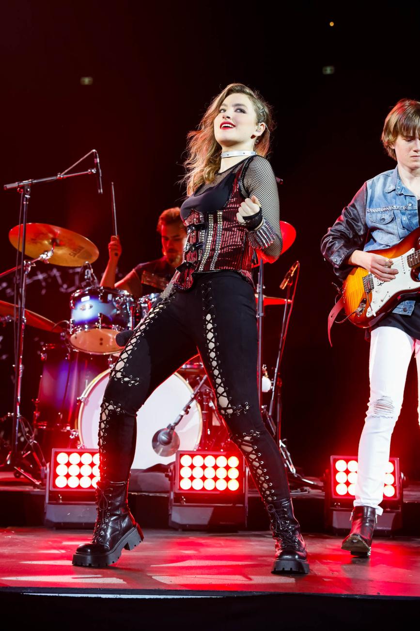 A singer fronting a band, wearing black boots, pants and top, and a red studded corset