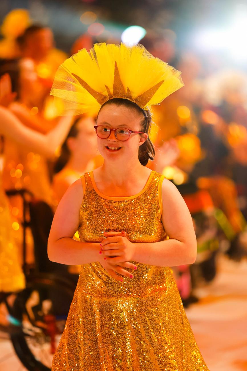 A young smiling performer wearing a sequined yellow dress, a tall, tulle headdress, and red glasses 