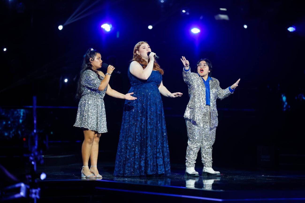 Three singers, wearing silver sequined dress and suit, and a long blue dress
