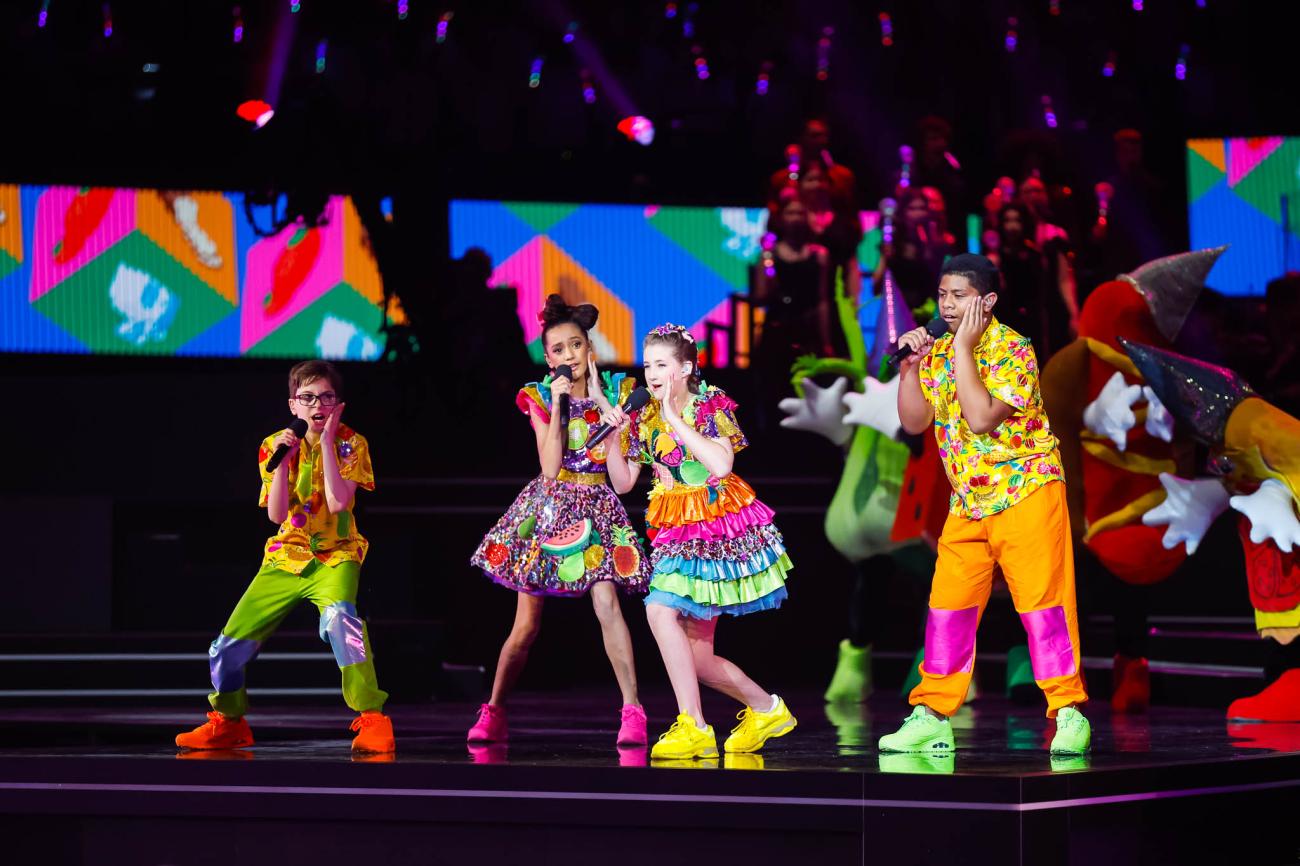 Four singers in brightly-coloured tropical themed costumes, with one hand on their cheeks