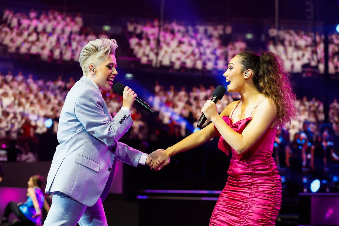 Two singers singing to each other, one wearing a grey suit and the other a deep pink ruffled dress a 