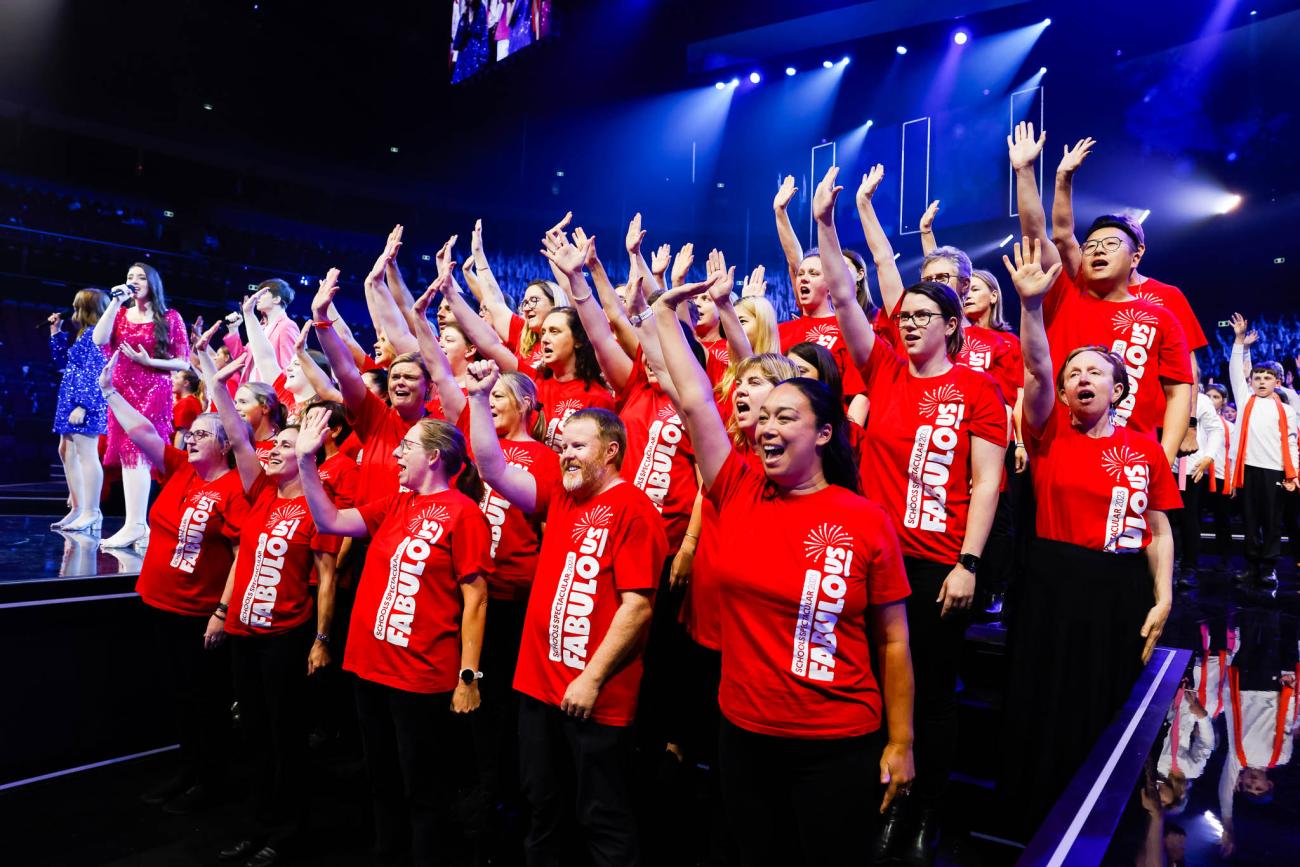 A group of adults wearing red shirts with the Schools Spectacular Fabulous watermark, with their right hands raised
