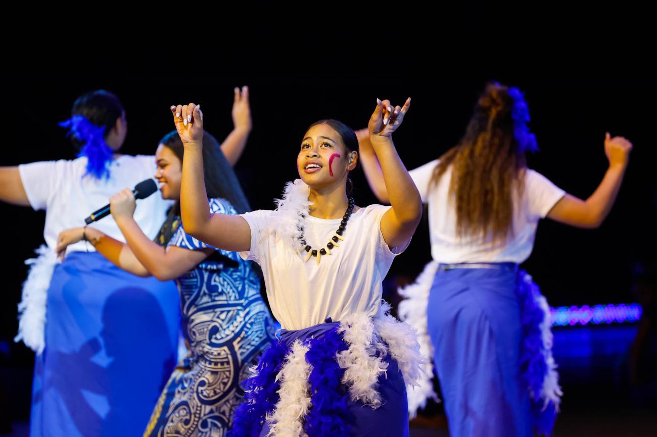 A group of islander dancer wearing white shirts and blue and white feathered dresses, with a singer wearing a blue and silver patterened dress