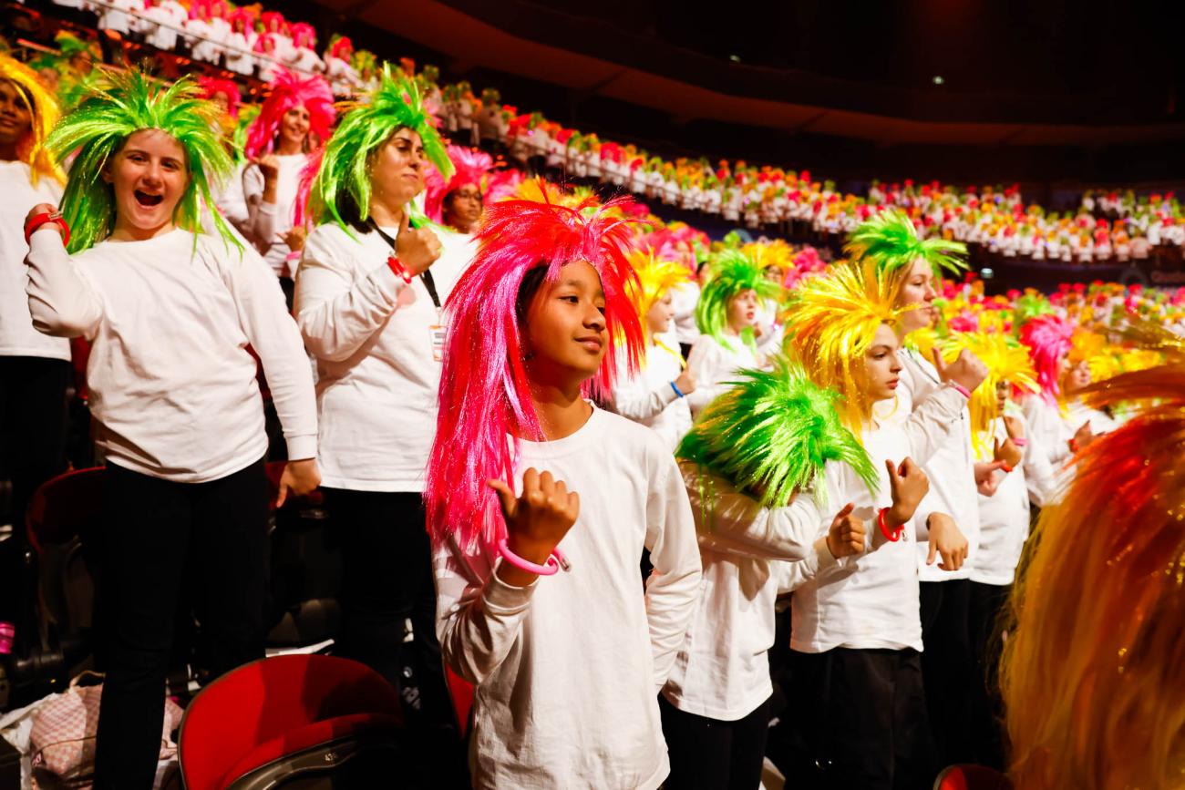 A large choir in the auditorium of an arena, each wearing white t-shirts and long, brightly coloured wigs.