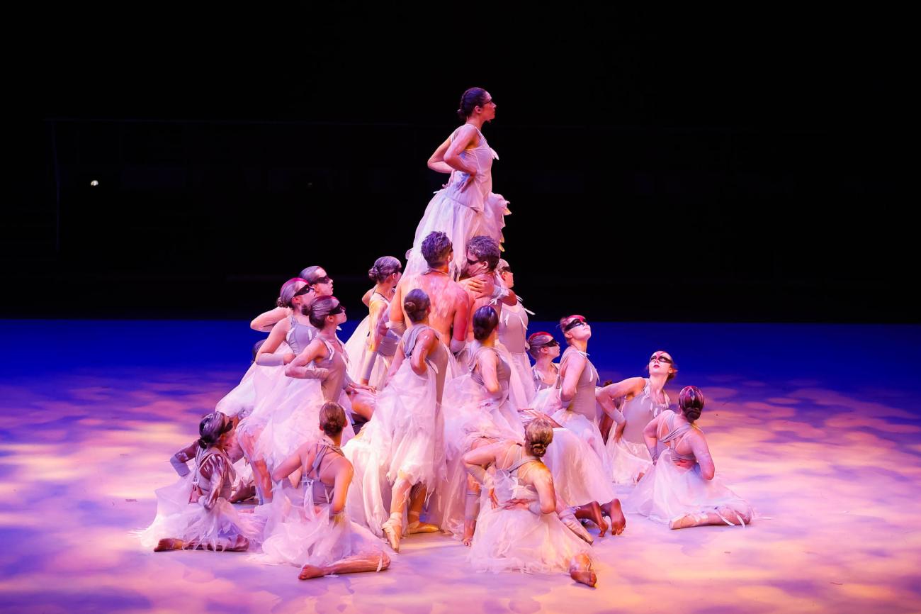 An ensemble of dancers in white and grey at different levels, some seated, others standing around an inner group of dancers lifting a female dancer above them. The dancers are in various bird-like poses, with bands of black and red paint on their faces