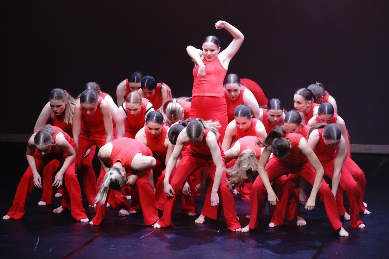 large clump of dancers in red bent over with one dancer in middle standing up with arms above head