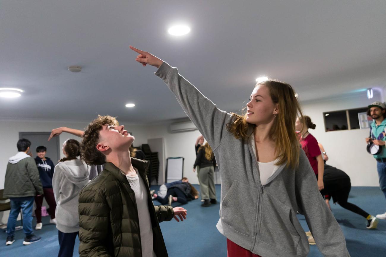 Two students do a warm up in a drama workshop with other students in the background.