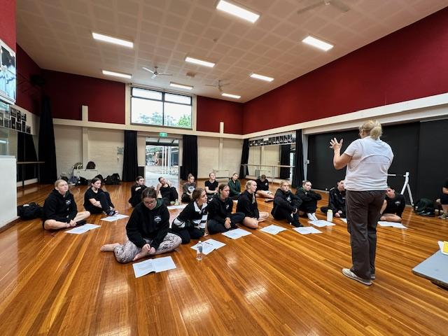 students seated in dance studio engaging with teacher standing at front of group.