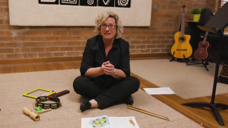 Donna Riles sitting on the floor surrounded by small musical instruments, a book of music and song lyrics.