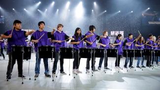 Drummers standing in a line, all wearing purple hooded tops and holding their drum sticks out in the same pose, each over their own drum.