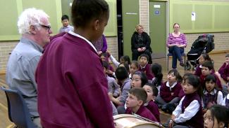Young music students sitting down and looking up to Richard Gill, and one student standing up next to a drum.