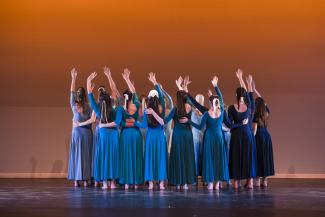 Dancers in blue dresses standing in a tight group on a stage facing an orange cyc, with one arm raised and the other across the back of a dancer standing next to them