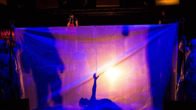 A scene from the NSW Public Schools Drama Company production Animal Farm with a silhouette of a person lying down with his arm reaching up