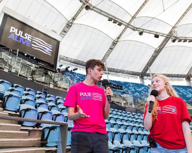Two students wearing Pulse Alive t-shirts standing in a stadium talking into and holding a microphone