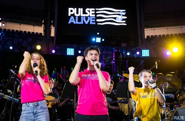 Three young students in pink and yellow Pulse Alive t-shirts, singing, with one raised fist