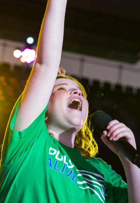 A student wearing a green t-shirt singing and looking up with one arm raised