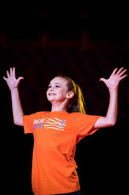 Smiling girl wearing orange Pulse Alive t-shirt with arms out and bent with hands up and spread
