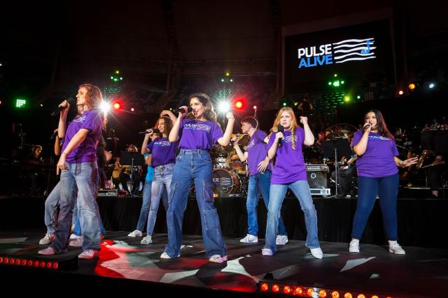 A group of singers wearing purple Pulse Alive t-shirt singing on stage in front of a band
