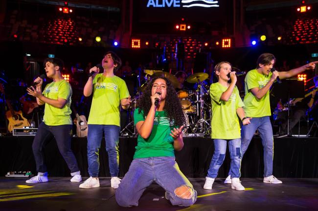 A group of young singers wearing green Pulse Alive t-shirt singing in front of a band