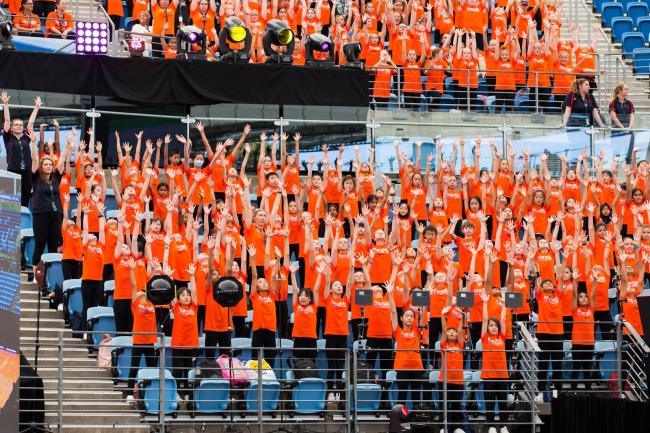 large group of students in a stadium stands, wearing orange t-shirts and with their arms above their heads