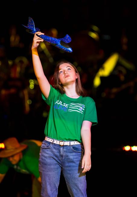 A girl wearing a green Pulse Alive t-shirt, holding a blue model plane above her head