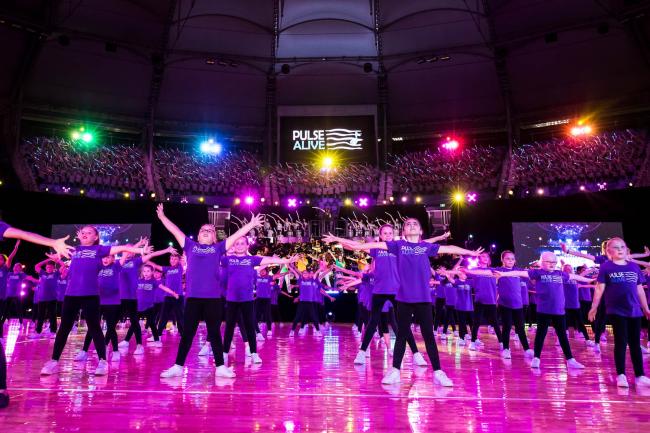 large group of students in a stadium, wearing purple t-shirts and with arms stretched out and legs akimbo