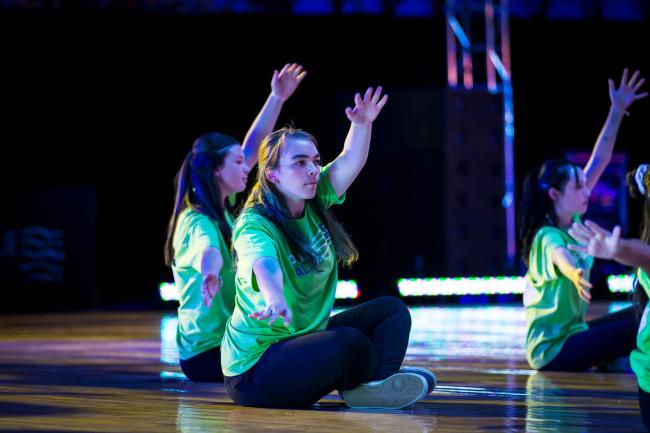 An ensemble of dancers wearing green Pulse Alive t-shirts, seated on the ground with one arm raised and other reaching out