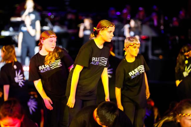 An ensemble of dancers wearing black Pulse Alive t-shirts, wearing headbands braided with the colours of the Aboriginal flag