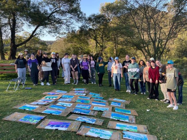 Students gather around the art work they have created in a grassy outdoor space.