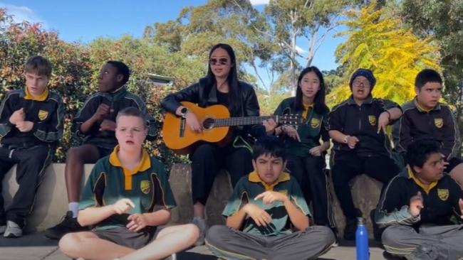 Eight students sit grouped around their teacher who is playing a guitar. They are singing and signing.