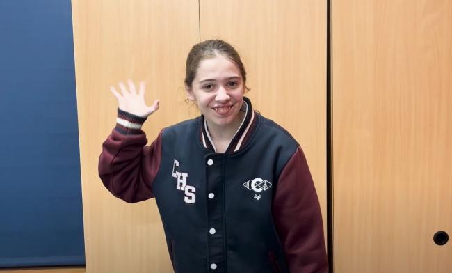 A female student waves at the camera while smiling. She wears a Crestwood High School jacket.