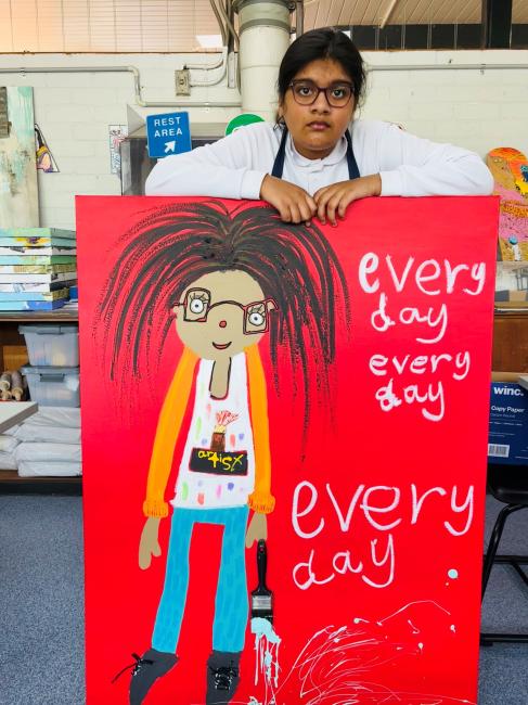 A student stands behind her large self-portrait. She has long frizzy hair and glasses, wearing blue pants, a white tank top and holding a paint brush. The background is bright red with the words ‘every day’ written 3 times in white.